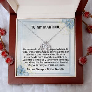 May include: A silver-toned necklace with a central knot-shaped pendant featuring a clear round stone. The necklace is displayed on a card with the text TO MY MARTINA, followed by a Spanish message, inside a dark brown wooden gift box.