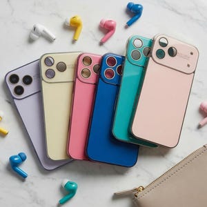 May include: A selection of brightly coloured phone cases in pastel shades of lilac, cream, pink, blue, and teal, with matching wireless earbuds. A small, beige zipped pouch is also present, all arranged on a marble surface.