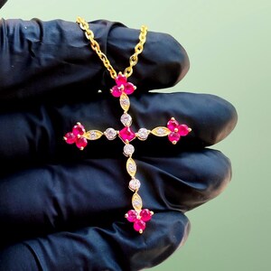 Puede incluir: Un collar con colgante de cruz de color dorado, con una cadena delicada. La cruz presenta piedras preciosas rosas vibrantes dispuestas en un patrón floral, intercaladas con piedras claras y brillantes. El collar se muestra sobre un fondo negro.