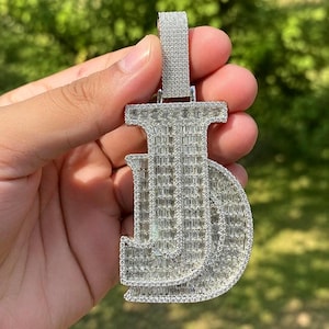 May include: A silver-toned pendant shaped like the letters JJ, fully encrusted with numerous small, clear, rectangular baguette-cut stones and a border of tiny round stones. The pendant features a rectangular bail at the top, also covered in stones.