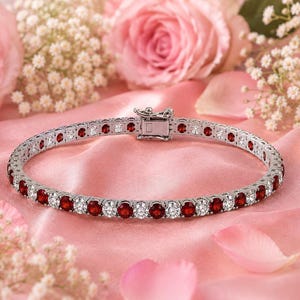 May include: A silver-toned tennis bracelet featuring an alternating pattern of round, faceted deep red stones and clear, sparkling white stones. The bracelet is secured with a rectangular metal box clasp mechanism at the centre of the circular band.