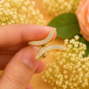 Puede incluir: Un anillo de serpiente en tono dorado con un diseño envolvente, adornado con piedras transparentes engastadas en pavé a lo largo del cuerpo y una pequeña piedra roja como ojo. La banda de metal pulido refleja la luz y muestra una cabeza de serpiente detallada.