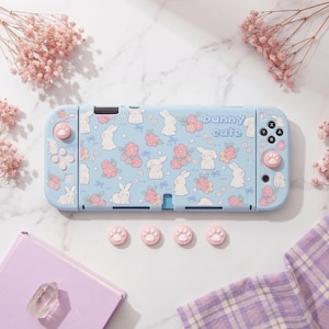 May include: A light blue Nintendo Switch Lite console with a bunny and floral pattern, and the text "bunny cute". Includes pink paw print thumb grips and a matching case. The console is surrounded by pink flowers and a purple book.