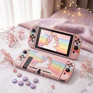 Puede incluir: Consola y funda Nintendo Switch rosas con diseño de unicornio y fondo arcoíris. La pantalla muestra una imagen de unicornio. Incluye tapas de pulgar moradas con diseños de luna y estrella.