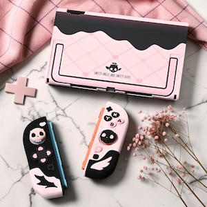 Può includere: Una base per console di gioco portatile e due controller con una combinazione di colori rosa e nero, motivi a cuore e ali. La base mostra il testo "SWEETY ANGEL AND SWEETY DEVIL" e un'icona a forma di teschio con ali e un'aureola.