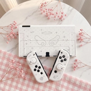 May include: A white video game console and two matching controllers featuring a light gray pattern of angel wings, a halo, stars, hearts, and musical notes. The controllers include white joystick covers with a swirl design and black buttons.