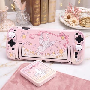 K&ouml;nnte beinhalten: Rosa Nintendo Switch Lite H&uuml;lle und passende Spielkassettenh&uuml;lle. Das Design zeigt eine Katze mit Fl&uuml;geln, Sterne und B&auml;nder. Die Kn&ouml;pfe sind wei&szlig; und schwarz.