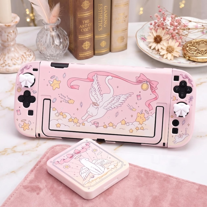 K&ouml;nnte beinhalten: Rosa Nintendo Switch Lite H&uuml;lle und passende Spielkassettenh&uuml;lle. Das Design zeigt eine Katze mit Fl&uuml;geln, Sterne und B&auml;nder. Die Kn&ouml;pfe sind wei&szlig; und schwarz.