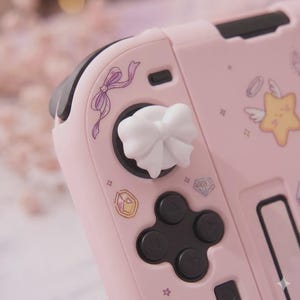 K&ouml;nnte beinhalten: Rosa Gaming-Controller mit wei&szlig;em, schleifenf&ouml;rmigem Joystick-Aufsatz. Der Controller hat schwarze Kn&ouml;pfe und ein Steuerkreuz. Verziert mit lila B&auml;ndern, Sternen und Edelstein-Grafiken. Ein s&uuml;&szlig;es Accessoire f&uuml;r Gamer.
