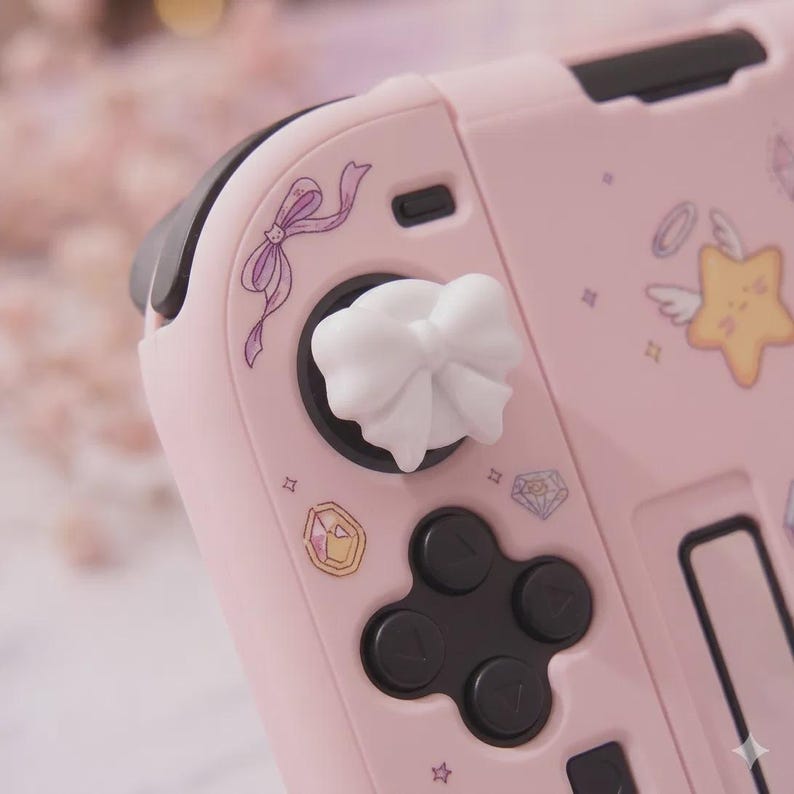 K&ouml;nnte beinhalten: Rosa Gaming-Controller mit wei&szlig;em, schleifenf&ouml;rmigem Joystick-Aufsatz. Der Controller hat schwarze Kn&ouml;pfe und ein Steuerkreuz. Verziert mit lila B&auml;ndern, Sternen und Edelstein-Grafiken. Ein s&uuml;&szlig;es Accessoire f&uuml;r Gamer.