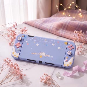 Puede incluir: Una funda lila para Nintendo Switch con un diseño de gato y estrellas. La funda tiene detalles rosas y blancos en los botones y bordes. El texto "WITH A CAT ALL IS WELL" está impreso en la funda.