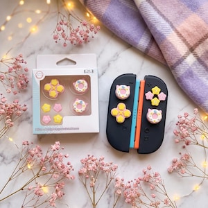 May include: A black Nintendo Switch controller with colourful button covers, including yellow cross-shaped buttons and pink donut-shaped buttons. A white box with matching button covers is also present, along with pink flowers and a plaid blanket.
