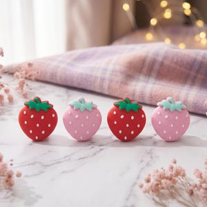 Op de afbeelding: Vier aardbeivormige decoraties. Twee zijn rood met groene toppen en witte stippen, en twee zijn roze met lichtblauwe toppen en witte stippen. Ze zijn gerangschikt op een wit marmeren oppervlak.