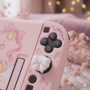 K&ouml;nnte beinhalten: Rosa Nintendo Switch Joy-Con-Controller mit wei&szlig;en, schleifenf&ouml;rmigen Daumengriffen. Der Controller hat dekorative Illustrationen von Schleifen, Monden und Sternen. Die Kn&ouml;pfe sind schwarz mit wei&szlig;en Buchstaben.