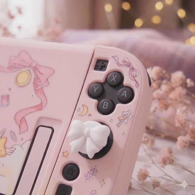 K&ouml;nnte beinhalten: Rosa Nintendo Switch Joy-Con-Controller mit wei&szlig;en, schleifenf&ouml;rmigen Daumengriffen. Der Controller hat dekorative Illustrationen von Schleifen, Monden und Sternen. Die Kn&ouml;pfe sind schwarz mit wei&szlig;en Buchstaben.