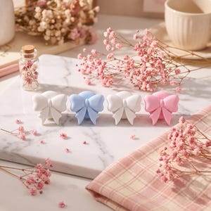 Può includere: Quattro pomelli decorativi per cassetti a forma di fiocco, disposti in fila. Il set include due pomelli bianchi, uno azzurro e uno rosa chiaro. Ogni pomello presenta una finitura opaca liscia e un design a fiocco dettagliato con un nodo.