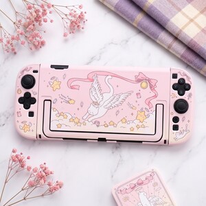 K&ouml;nnte beinhalten: Rosa Nintendo Switch-Konsole mit individuellem Design mit einer gefl&uuml;gelten Katze, Sternen und B&auml;ndern. Das Design ist im Cartoon-Stil gehalten. Die Konsole hat schwarze Steuerkn&uuml;ppel und Tasten. Eine passende H&uuml;lle ist ebenfalls sichtbar.