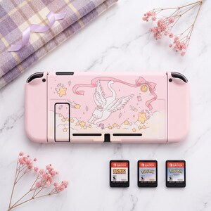 K&ouml;nnte beinhalten: Eine rosa Nintendo Switch-Konsole mit einem dekorativen Design mit einer gefl&uuml;gelten Katze, Sternen und B&auml;ndern. Drei Spielkassetten sind unter der Konsole sichtbar. Ein gefaltetes kariertes Tuch und rosa Blumen befinden sich im Hintergrund.