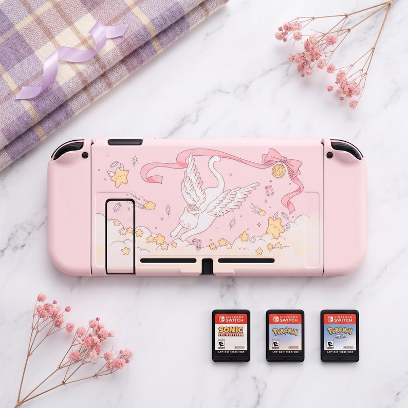 K&ouml;nnte beinhalten: Eine rosa Nintendo Switch-Konsole mit einem dekorativen Design mit einer gefl&uuml;gelten Katze, Sternen und B&auml;ndern. Drei Spielkassetten sind unter der Konsole sichtbar. Ein gefaltetes kariertes Tuch und rosa Blumen befinden sich im Hintergrund.