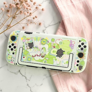 May include: A light green gaming console protective case featuring a collage of cartoon stickers including black cats, pandas, clovers, musical notes, and food items. The case has a black frame outline and cutouts for buttons and joysticks.