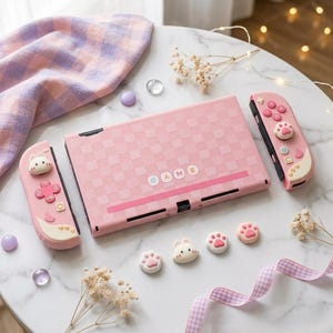 Könnte beinhalten: Rosa Nintendo Switch Konsole und Controller mit Schachbrettmuster. Die Controller haben Knöpfe in Hasen- und Pfotenform. Auf der Konsole steht das Wort „GAME“. Zubehör umfasst Pfoten- und Hasen-Charms.