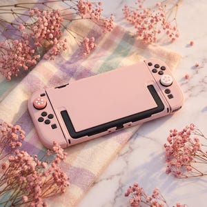 May include: A handheld gaming console featuring a solid pastel pink protective case. The device includes two detachable controllers with matching pink thumbstick caps, each decorated with a small white cherry blossom flower graphic.