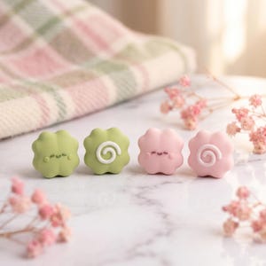 May include: Four small, flower-shaped charms arranged in a row. Two are light green and two are light pink. One of each colour features a simple, smiling face, while the other two feature a raised white spiral design on their front surfaces.