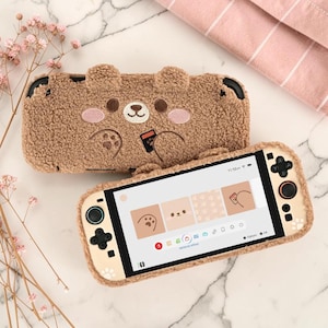 May include: A brown, fuzzy, plush protective case for a handheld gaming console, featuring an embroidered bear face with pink cheeks and paws. A matching gaming console with a cream-colored skin and paw print accents is displayed in front of the case.
