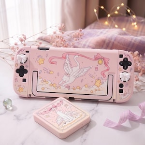 Peut inclure: Une console Nintendo Switch rose et un étui assorti, décorés d'un motif fantaisiste. Le motif comprend un chat ailé, des étoiles et des rubans. La console a des capuchons de joystick blancs et des boutons noirs.