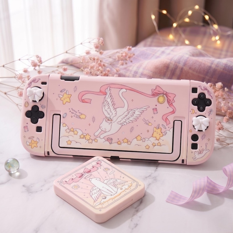 K&ouml;nnte beinhalten: Eine rosa Nintendo Switch-Konsole und eine passende H&uuml;lle mit einem fantasievollen Design. Das Design zeigt eine Katze mit Fl&uuml;geln, Sterne und B&auml;nder. Die Konsole hat wei&szlig;e Joystick-Kappen und schwarze Kn&ouml;pfe.