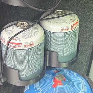 Puede incluir: Dos bidones de combustible Coleman de propano y butano, asegurados en un soporte negro. Los bidones son blancos y grises con información del producto impresa. Un recipiente azul con una válvula roja es visible debajo.