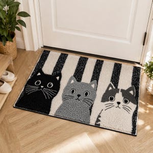 Könnte beinhalten: Rechteckige Fußmatte mit drei Katzenköpfen. Die Katzen sind schwarz, grau und weiß, vor einem Hintergrund aus schwarz-weißen vertikalen Streifen. Die Matte liegt vor einer weißen Tür.