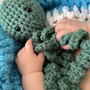 May include: A green crocheted octopus toy with large black eyes. The octopus is being held by a baby's hand.