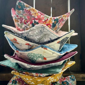 May include: A stack of seven fabric bowls in various colors and patterns. The bowls are made of quilted fabric and have a star-shaped design. The bowls are stacked in a pyramid shape, with the largest bowl at the bottom and the smallest bowl at the top.