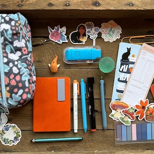 May include: A collection of stationery and accessories in a wooden box. Includes a floral pencil case, stickers, a notebook, pens, a bookmark, and a heart-shaped object. The items are arranged on a wooden surface.