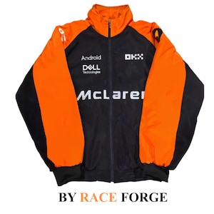 Può includere: Giacca da corsa nera e arancione con il logo McLaren e i loghi degli sponsor. La giacca ha una zip completa e un cappuccio. Il testo "BY RACE FORGE" è stampato in basso.