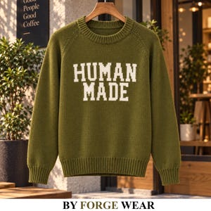 May include: An olive green long-sleeve knit jumper featuring a crew neckline and ribbed cuffs and hem. The front of the jumper displays the text HUMAN MADE in large, bold, white block letters centred across the chest area of the garment.