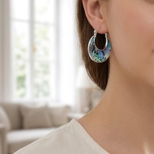 May include: Silver hoop earrings with a crescent-shaped abalone shell inlay. The iridescent shell features shades of blue, green, and silver. The earrings are shown on a person's ear, with a soft background.