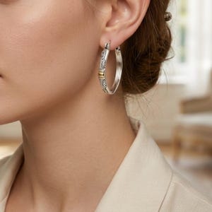 May include: Silver hoop earrings with ornate detailing and a gold-colored accent. The earrings are worn on a person's ear, showcasing their design and craftsmanship. The earrings are a classic jewelry piece.