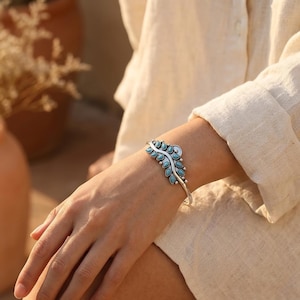 May include: A silver-toned cuff bracelet with a leaf design. The bracelet features turquoise-colored stones set in a detailed pattern. The bracelet is worn on a wrist, showcasing its intricate design and craftsmanship.