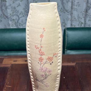 May include: Cream-colored ceramic vase with a crackled finish. The vase features a floral design in pink and purple hues. The vase has a tall, slender shape with a series of decorative holes along the sides.