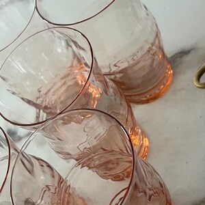 May include: A collection of six pink-tinted, clear glass tumblers. The glasses have a unique, curved shape with a textured design on the lower half. The rims are outlined with a darker pink hue. The glasses are arranged on a light-colored surface.