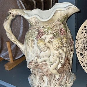 May include: A cream-colored ceramic pitcher with a detailed relief design. The pitcher features cherubic figures, grape vines, and a textured surface. The handle is intricately sculpted, and the rim has a scalloped edge. A decorative piece for the home.