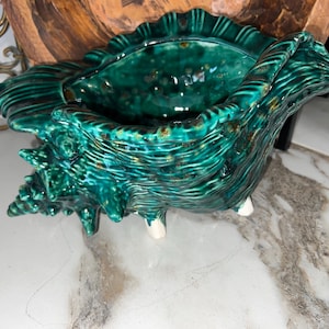 May include: A ceramic seashell-shaped planter in shades of teal and green. The planter has a textured surface with a glossy finish and sits on three small white feet. The interior is a darker teal, and the edges are ruffled.