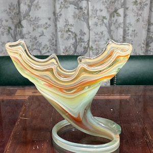 May include: A decorative glass bowl with a swirling design in shades of yellow, orange, and green. The bowl has a unique shape with a wide, open top and a curved base. The base is supported by a circular ring.