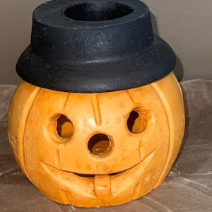 May include: A ceramic Halloween pumpkin with a smiling face and a black hat. The pumpkin has cut-out eyes and a mouth, and is painted in a bright orange color. A festive decoration for the fall season.