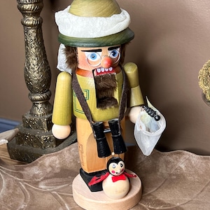 May include: A wooden nutcracker figure dressed as an explorer, wearing a green pith helmet with a white fabric band, round glasses, and a brown strap holding black binoculars. It carries a small white mesh net and stands on a round base.
