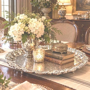 May include: A large, round, silver-toned metal serving tray with an ornate, scalloped edge and intricate embossed detailing. The tray holds a mercury glass vase filled with white flowers, a lit votive candle, two stacked vintage books, and a small box.