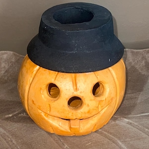 May include: A carved orange pumpkin with a black hat, featuring three eye holes and a carved smiling mouth. The Halloween decoration is set on a textured, brown surface.