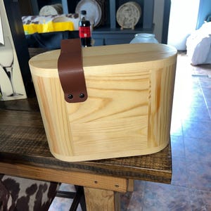 May include: A light-colored wooden picnic basket with a brown leather handle. The basket has a rectangular shape with rounded edges. The wood grain is visible. The handle is attached with two black rivets. The basket is sitting on a wooden table.